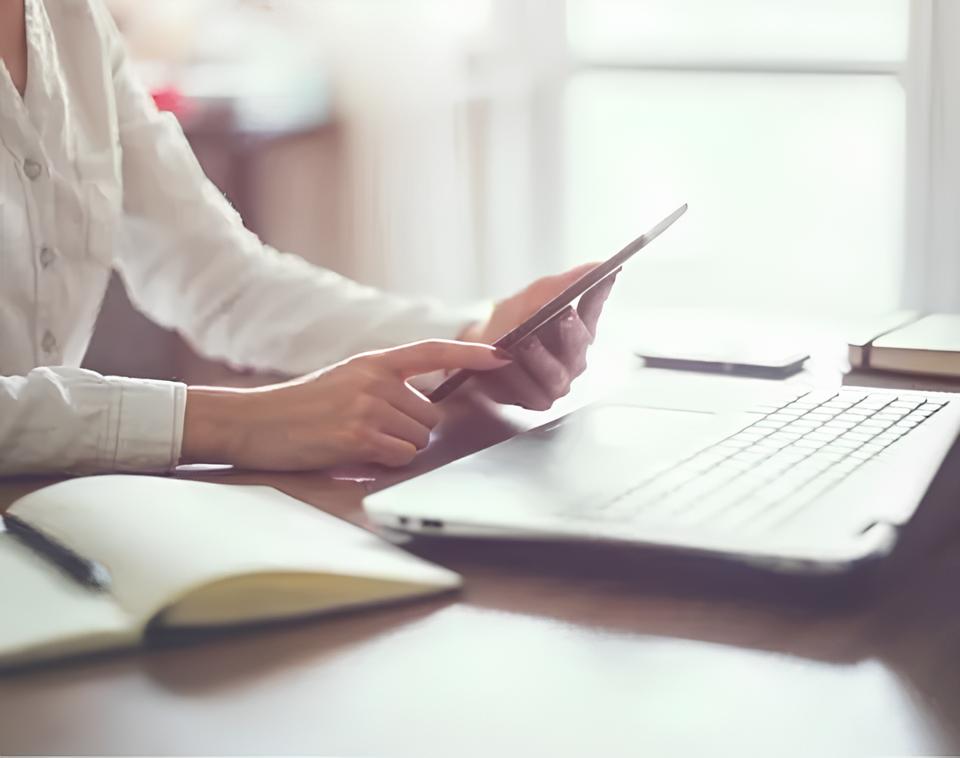 Eine Person, die ein Tablet benutzt, während sie an einem Schreibtisch mit einem Laptop, einem Notebook und einem Smartphone sitzt, in einer Umgebung mit weichem, natürlichem Licht.