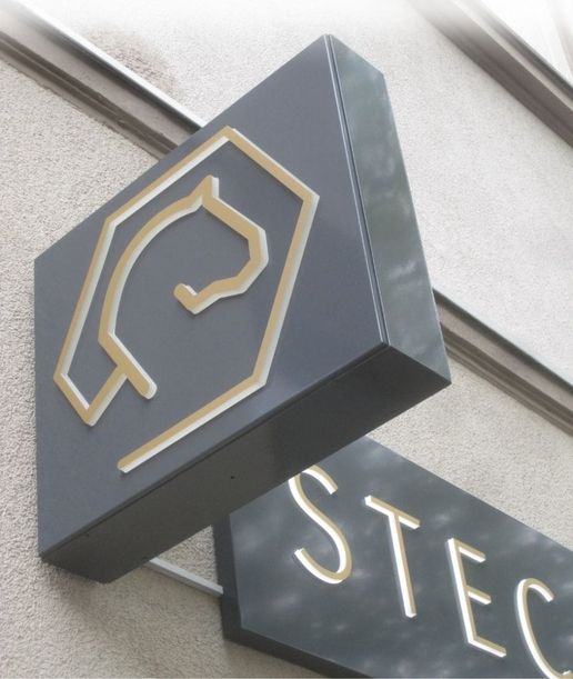 Schild mit einem minimalistischen Pferdelogo in Gold und dem Text STEIG in schlichten, modernen Buchstaben auf grauem Hintergrund.