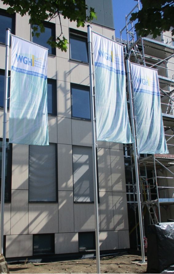 Drei Banner mit dem Schriftzug "WGH" hängen vor einem modernen, im Bau befindlichen Gebäude, im Hintergrund sind Gerüste zu sehen; ein Kran hebt eine große Glasplatte an, während Arbeiter auf einer Plattform bei der Montage an einer Gebäudefassade unter bewölktem Himmel helfen.