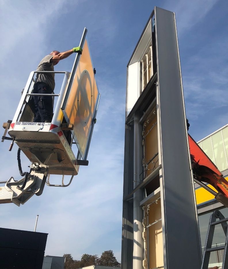Ein Arbeiter in einem Aufzug ersetzt ein großes Schild an einem hohen Gebäude unter blauem Himmel, während ein Baufahrzeug in der Nähe steht.