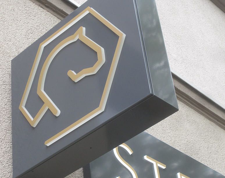 Schild mit einem minimalistischen Pferdelogo in Gold und dem Text STEIG in schlichten, modernen Buchstaben auf grauem Hintergrund.
