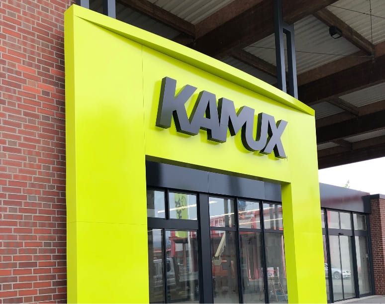 Leuchtend gelbes Schild mit dem Namen "KAMUX" an einer modernen Schaufensterfront, eingerahmt von rotem Backstein und großen Glastüren.
